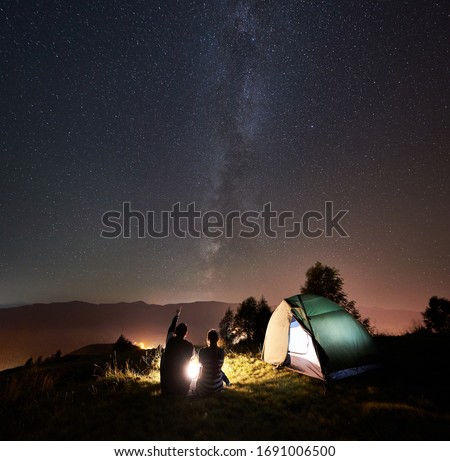 Back view of young couple tourists having a rest at campfire near glowing tent under night sky full of stars and Milky way. On the background beautiful starry sky, mountains and luminous town