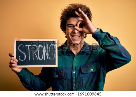 Young handsome man holding blackboard with strong word over isolated yellow background with happy face smiling doing ok sign with hand on eye looking through fingers