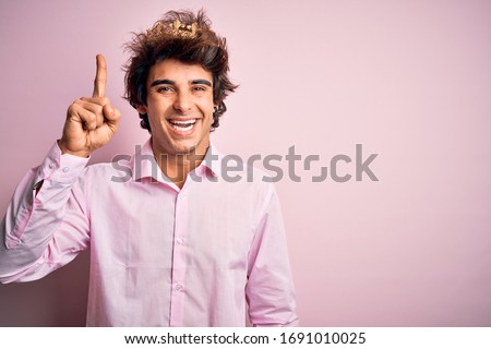 Young handsome man wearing king crown standing over isolated pink background pointing finger up with successful idea. Exited and happy. Number one.