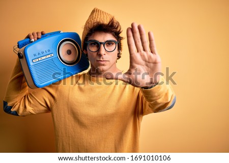Young handsome man holding vintage radio standing over isolated yellow background with open hand doing stop sign with serious and confident expression, defense gesture