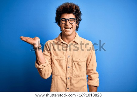 Young handsome man wearing casual shirt standing over isolated blue background smiling cheerful presenting and pointing with palm of hand looking at the camera.