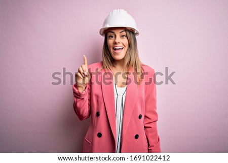 Young beautiful brunette architect woman wearing safety helmet over pink background pointing finger up with successful idea. Exited and happy. Number one.