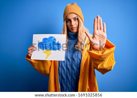 Young blonde woman wearing raincoat for rainy weather holding paper with clound and thunder with open hand doing stop sign with serious and confident expression, defense gesture