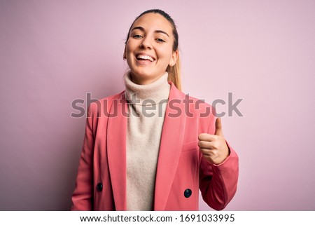 Young beautiful blonde businesswoman with blue eyes wearing elegant pink jacket doing happy thumbs up gesture with hand. Approving expression looking at the camera showing success.