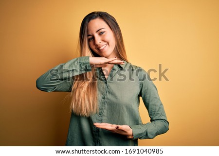 Young beautiful blonde woman with blue eyes wearing green shirt over yellow background gesturing with hands showing big and large size sign, measure symbol. Smiling looking at the camera. Measuring