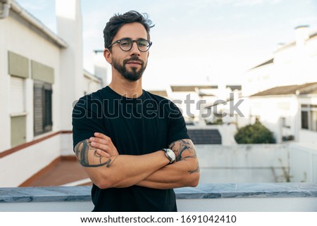 Confident stylish guy with tattoos posing on apartment balcony or terrace. Young man in glasses standing outside with arms crossed and looking at camera. Male portrait concept Royalty-Free Stock Photo #1691042410