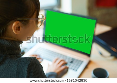 Back view woman freelancer in eyeglasses sitting at desk and typing on laptop with a green screen on monitor. Concept remote work, freelance, using laptop computer or net-book.