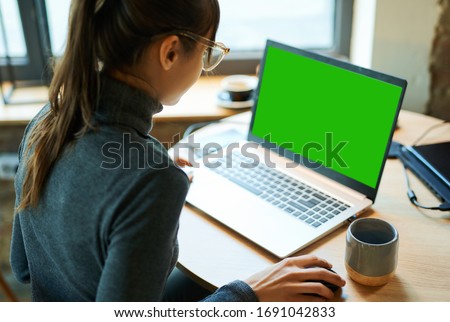 Rear view woman freelancer in eyeglasses working in cafe, using laptop computer with a green screen on monitor. Concept remote work, freelance, working on laptop computer or net-book.