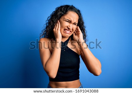 Young beautiful woman with curly hair wearing casual t-shirt standing over blue background covering ears with fingers with annoyed expression for the noise of loud music. Deaf concept.