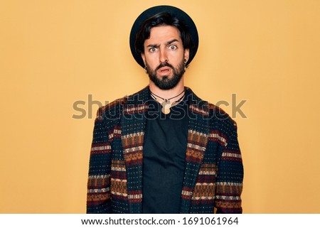 Young handsome hispanic bohemian man wearing hippie style and boho hat In shock face, looking skeptical and sarcastic, surprised with open mouth