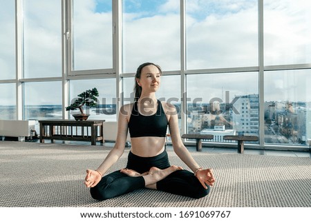 Young athletic girl meditating in the Lotus position in the white yoga studio with panoramic windows overlooking the city.Concept of healthy lifestyle.