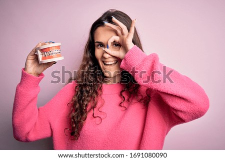 Beautiful woman with curly hair holding plastic teeth with dental braces over pink background with happy face smiling doing ok sign with hand on eye looking through fingers