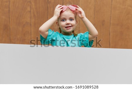 Little smiling child girl heart shaped glasses standing behind a white blank panel against wooden background. Funny face. Peeking out from behind a banner, empty space for text.