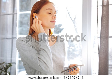 Long-haired girl near the window. Blonde with a wireless earphone in her ear. Woman in a light gray blouse. Girl with a phone in her hands.