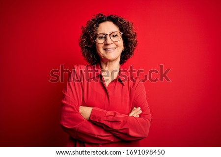Middle age beautiful curly hair woman wearing casual shirt and glasses over red background happy face smiling with crossed arms looking at the camera. Positive person.