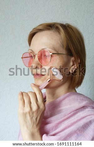 Mature woman using pink face roller. Skin treatment. Vertical photo.