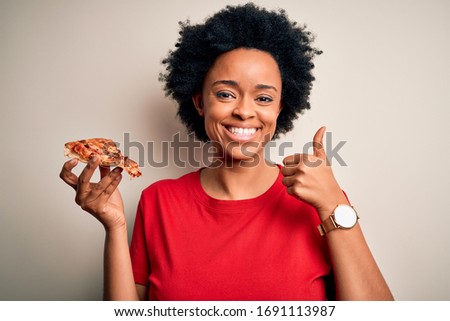 Young African American afro woman with curly hair eating slice of delicious Italian pizza happy with big smile doing ok sign, thumb up with fingers, excellent sign