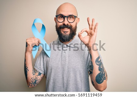 Handsome bald man with beard and tattoos holding blue cancer ribbon over isolated background doing ok sign with fingers, excellent symbol