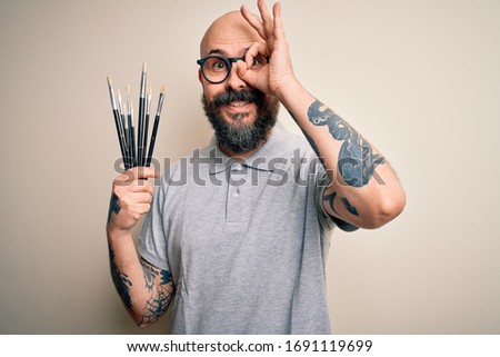 Handsome bald artist man with beard and tattoo painting using painter brushes with happy face smiling doing ok sign with hand on eye looking through fingers