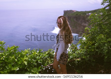Beautiful girl traveler with long hair and a slender body stands on a cliff of a cliff overlooking the waves of the blue ocean.Bali Island, Indonesia. Travel and tourism concept in exotic and tropical