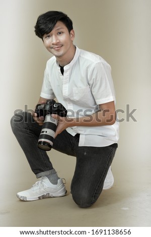 Asian man in white shirt kneeling while holding digital camera in hands. Male photographer looking at camera smiling. Isolated plain background copy space.