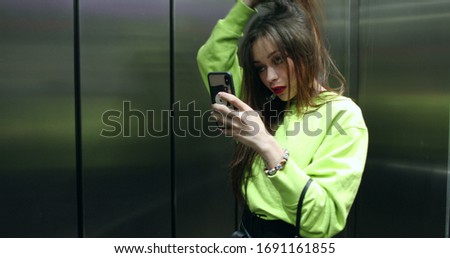 
Pretty girl adjusting hair taking photo in front of mirror at elevator. Attractive young woman posing for picture taken with phone, adjusting hair