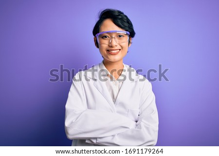 Young beautiful asian scientist girl wearing coat and glasses over purple background happy face smiling with crossed arms looking at the camera. Positive person. Royalty-Free Stock Photo #1691179264