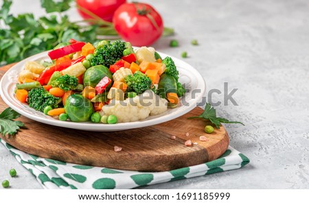 Mix of stewed vegetables in a ceramic plate on the served table. Boiled Brussels sprouts, carrots, broccoli, peas, peppers and corn. Cooked vegan or vegetarian food. Royalty-Free Stock Photo #1691185999
