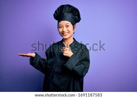 Young beautiful chinese chef woman wearing cooker uniform and hat over purple background Showing palm hand and doing ok gesture with thumbs up, smiling happy and cheerful