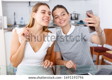Two smiling young girls sitting at kitchen table and taking selfie