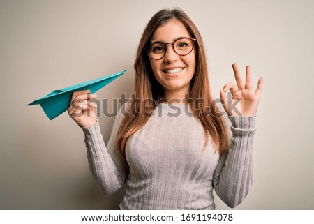 Young woman wearing glasses holding orimagi paper plane over isolated background doing ok sign with fingers, excellent symbol
