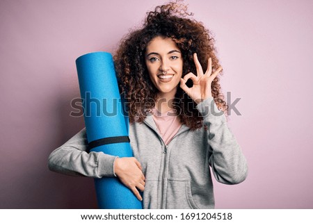 Young beautiful sporty woman with curly hair and piercing holding mat doing yoga doing ok sign with fingers, excellent symbol