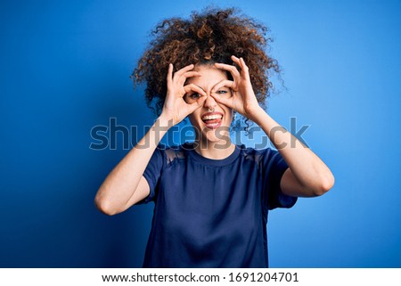 Young beautiful woman with curly hair and piercing wearing casual blue t-shirt doing ok gesture like binoculars sticking tongue out, eyes looking through fingers. Crazy expression.