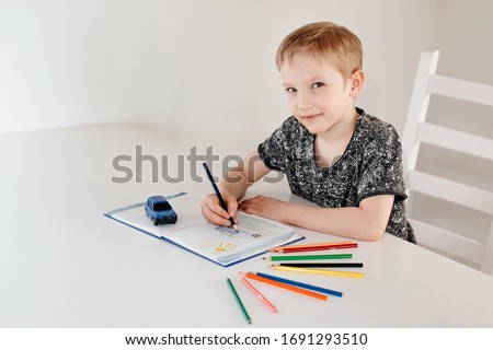 a  blond-haired 8-year-old boy sits at a table with a pencil in his hands and looks at the camera. child draws with colored pencils sitting at a white table on a light background