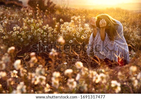 A bent down young girl with long blond hair and her hands looking up in a woolen grey poncho on a field with white flowers during sunset, with the sun on her back. Royalty-Free Stock Photo #1691317477