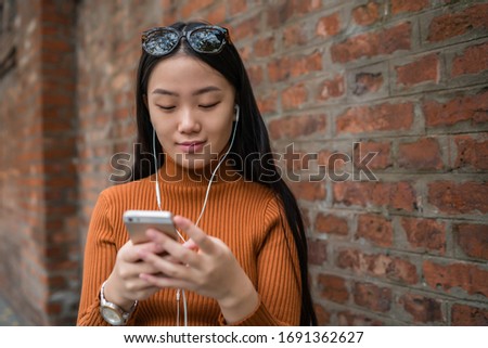 Portrait of young Asian woman using her mobile phone with earphones outdoors in the street. Urban and communication concept.