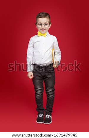 Positive little kid in trendy outfit with bow tie and spectacles looking at camera while standing with book in hand against red background