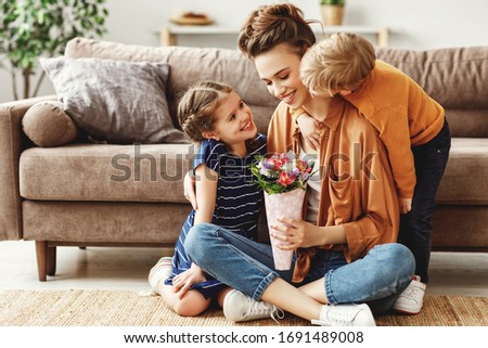 Joyful relaxed mother sitting on floor near sofa and enjoying presented bouquet of colorful fresh flowers while small daughter and son hugging and congratulating with Mother Day holiday at home
