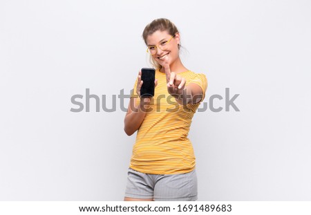 young pretty latin woman smiling proudly and confidently making number one pose triumphantly, feeling like a leader with a smartphone against white wall