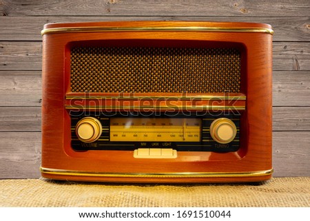 A red-brown old radio made of wood. There are knobs. The scale shows medium wave and FM. The radio stands on a fabric ceiling in front of a wooden wall.