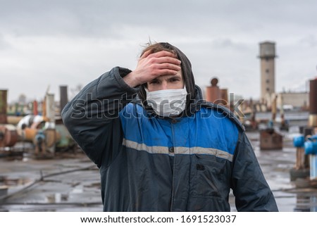 factory worker does not feel well at the workplace. Disposable protective bandage on the face. symptoms of coronavirus. holding his hand to his head. headache. virus protection Royalty-Free Stock Photo #1691523037