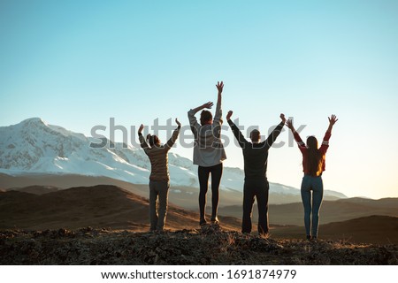 Four happy friends are standing with raised arms and looking at sunset mountains and sky. Unrecognized peoples