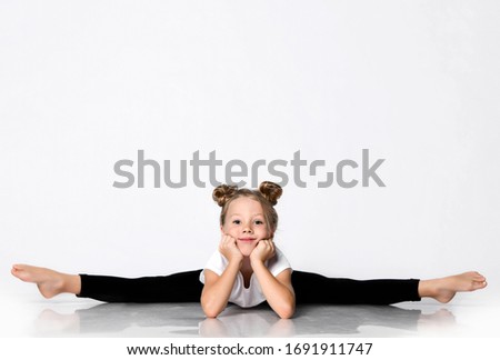 Cheerful athletic blonde kid girl does gymnastic exercises stretching at home in studio splits bend forward with her head leaning her hands over white background