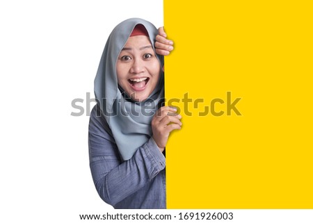 Young Asian muslim businesswoman smiling and peeking behind blank white advertising billboard wign board with copy space