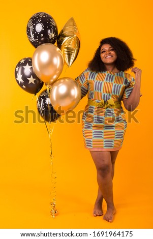 Holidays, birthday party and fun concept - Portrait of smiling young African-American young woman looking sweet on yellow background holding balloons.
