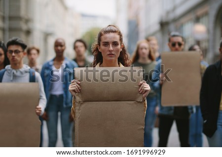 Female activist with a blank banner protesting on the street with protestors in background. People doing demonstration on the street holding sign boards.