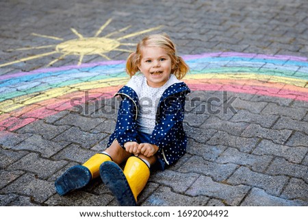 Happy little toddler girl in rubber boots with rainbow painted with colorful chalks on ground during pandemic coronavirus quarantine. Children painting rainbows along with the words Let's all be well