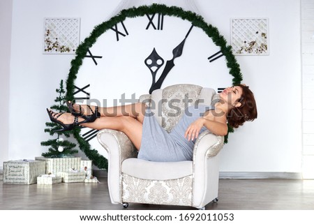 Young brunette woman, wearing grey dress, sitting lying resting relaxing on white beige armchair in light room with big clock on background. Lifestyle photo shoot in apartment. New year celebration