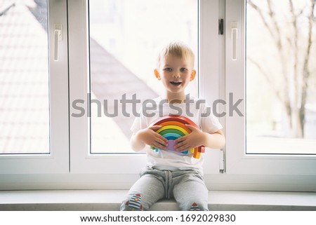 Child boy on background of window holds wooden rainbow toy. Photo of kids leisure and daycare at home. Concept of positive visual support during quarantine Pandemic Coronavirus Covid-19 at home.