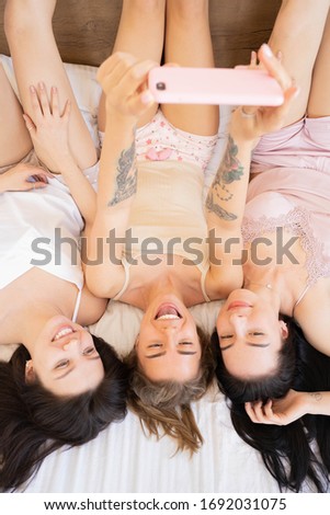 Three pretty young girls taking selfie while lying on the bed at bachelorette party. Top view of bride taking selfie with bridesmaids making funny faces. Home isolated, stay at home concept